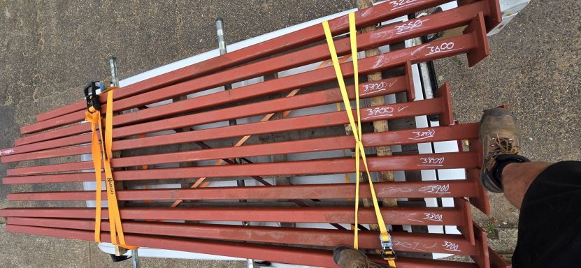 Red primed structural steel beams loaded on a trailer for delivery, Norwich Stack of red oxide primed steel posts with welded base plates, chalk length marks, strapped for delivery