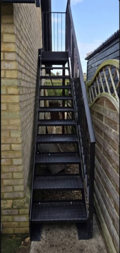 Black metal outdoor staircase leading up alongside a brick wall and wooden fence.